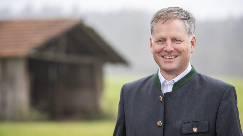 Dr. Rudolf Neumaier im Porträt. Er trägt einen Trachtenjanker. Im Hintergrund undcharf ein Feld und ein Feldstadl.