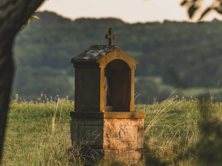 Ein steinerner Bildstock auf einer Wiese im Abendlicht. Im Hintergrund sind Hügel zu sehen.