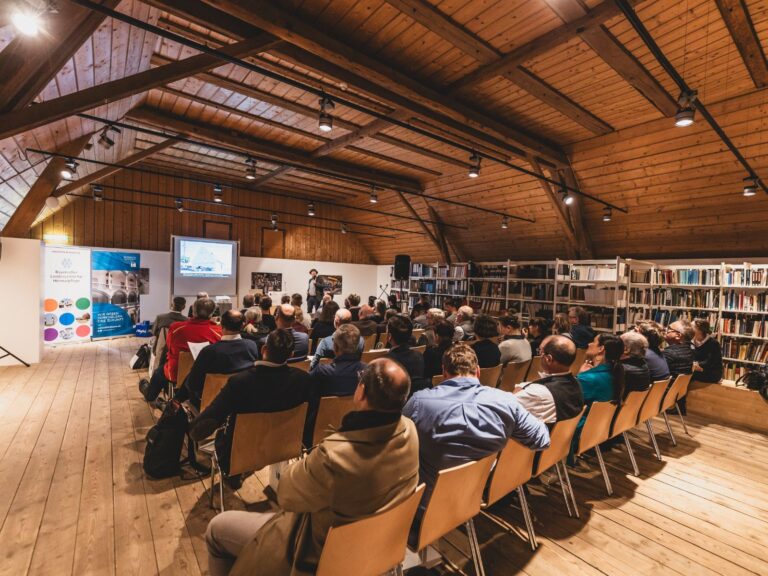 Publikum von hinten aufgenommen, sitzend bei einer Tagung, vorne steht Vinzenz Dufter und hält eine Powerpoint-Präsentation. Die Tagung findet in einem großen Holzgebäude statt, an einer Wand stehen Bücherregale.